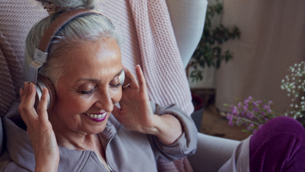 A relaxed senior woman sitting in armchair with headphones and listening to music indoors at home.