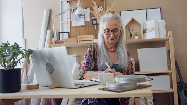 A creative senior woman with laptop sitting by desk and working by laptop in office.