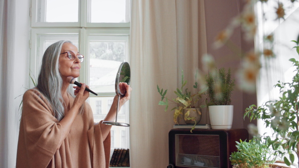 A senior woman looking at mirror and applying make up at home, selfcare concept.