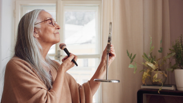 A senior woman looking at mirror and applying make up at home, selfcare concept.