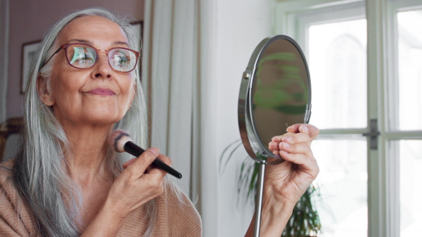 A senior woman looking at mirror and applying make up at home, selfcare concept.