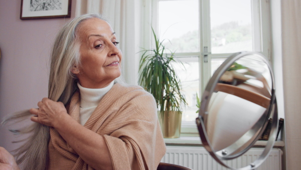 A senior woman looking at mirror and styling hair at home, selfcare concept.