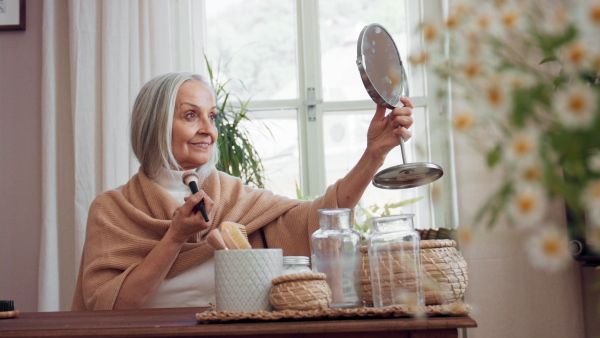 A senior woman looking at mirror and applying make up at home, selfcare concept.