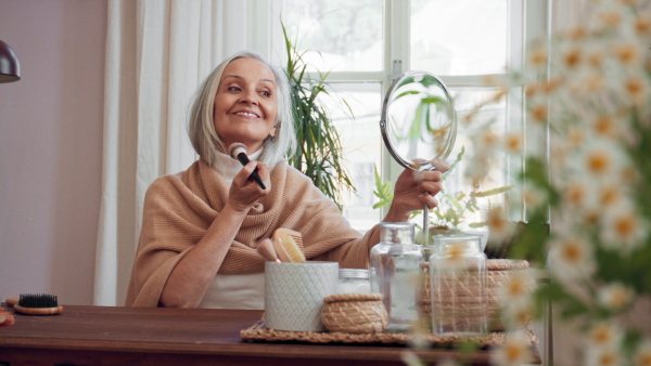 A senior woman looking at mirror and applying make up at home, selfcare concept.