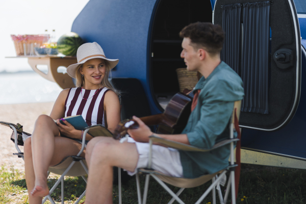 Happy young couple sitting together in front of van, camping and playing at a guitair.