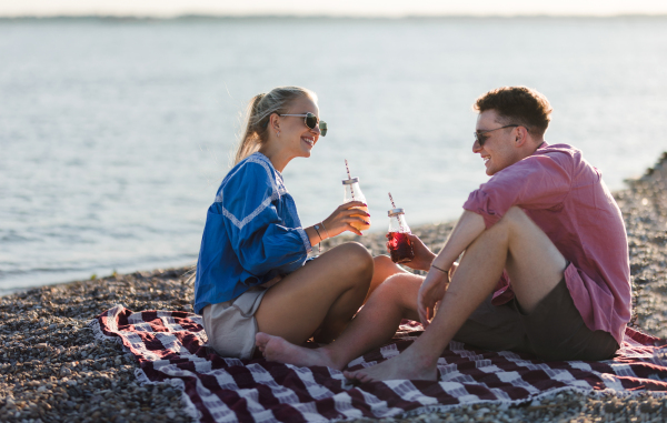 Happy young couple dating together in beach, sitting on the blanket and having toast with bottled beer. Enjoying holiday time together.