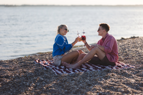 Happy young couple dating together in beach, sitting on the blanket and having toast with bottled beer. Enjoying holiday time together.