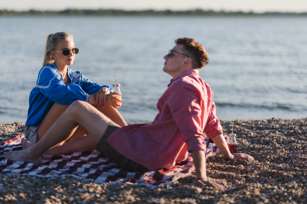 Happy young couple dating together in beach, sitting on the blanket and having toast with bottled beer. Enjoying holiday time together.