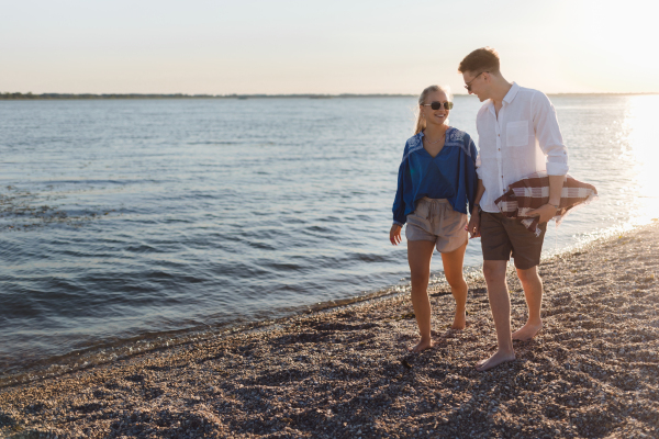 Happy young couple with blanket walking in the beach during sunset, prepairing for a picnic.