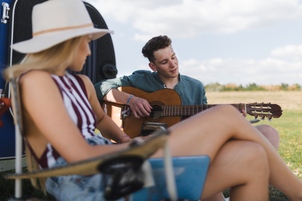 Happy young couple sitting together in front of van, camping and playing at a guitair.