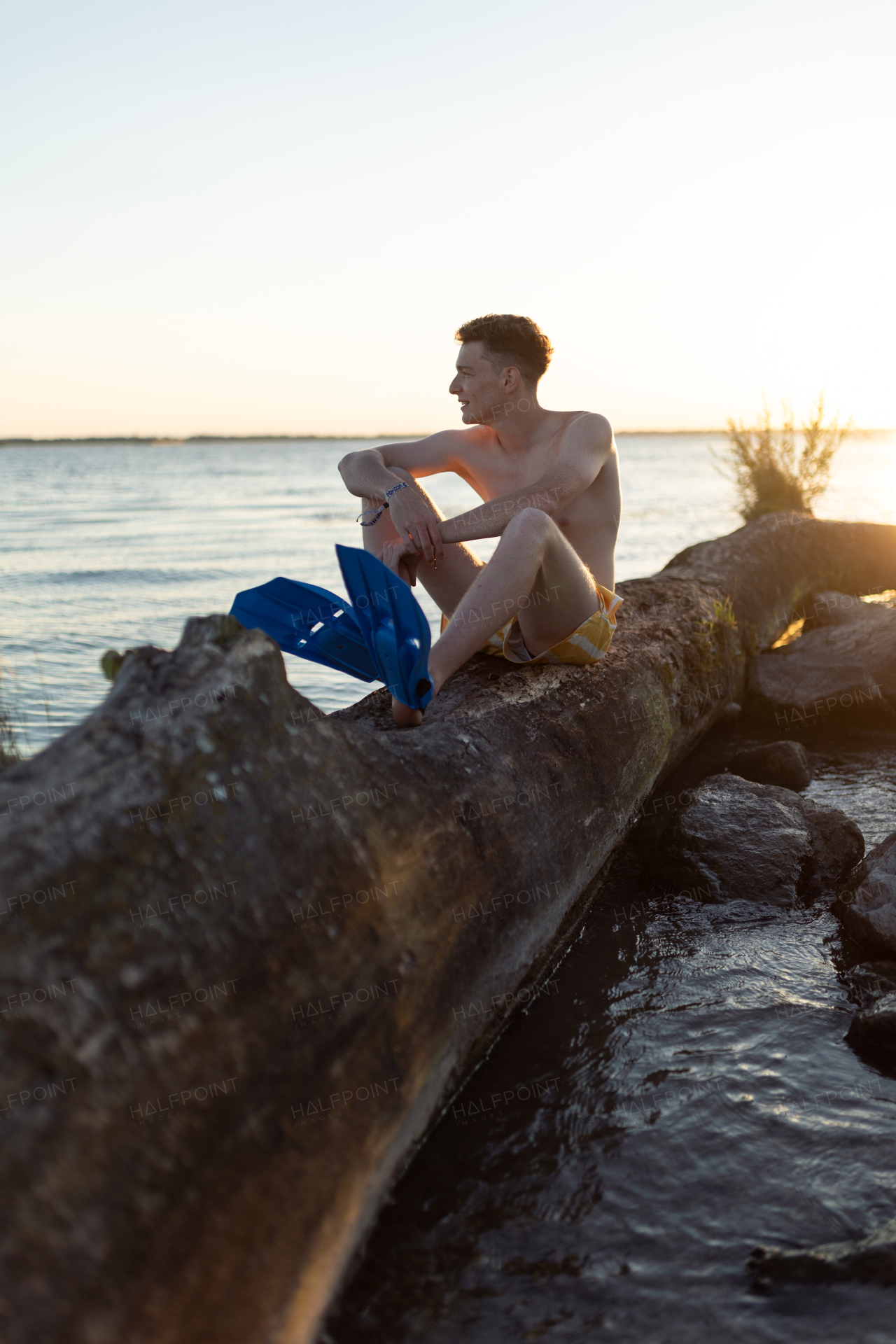 Young man sitting on tree stump in lake during sunset.