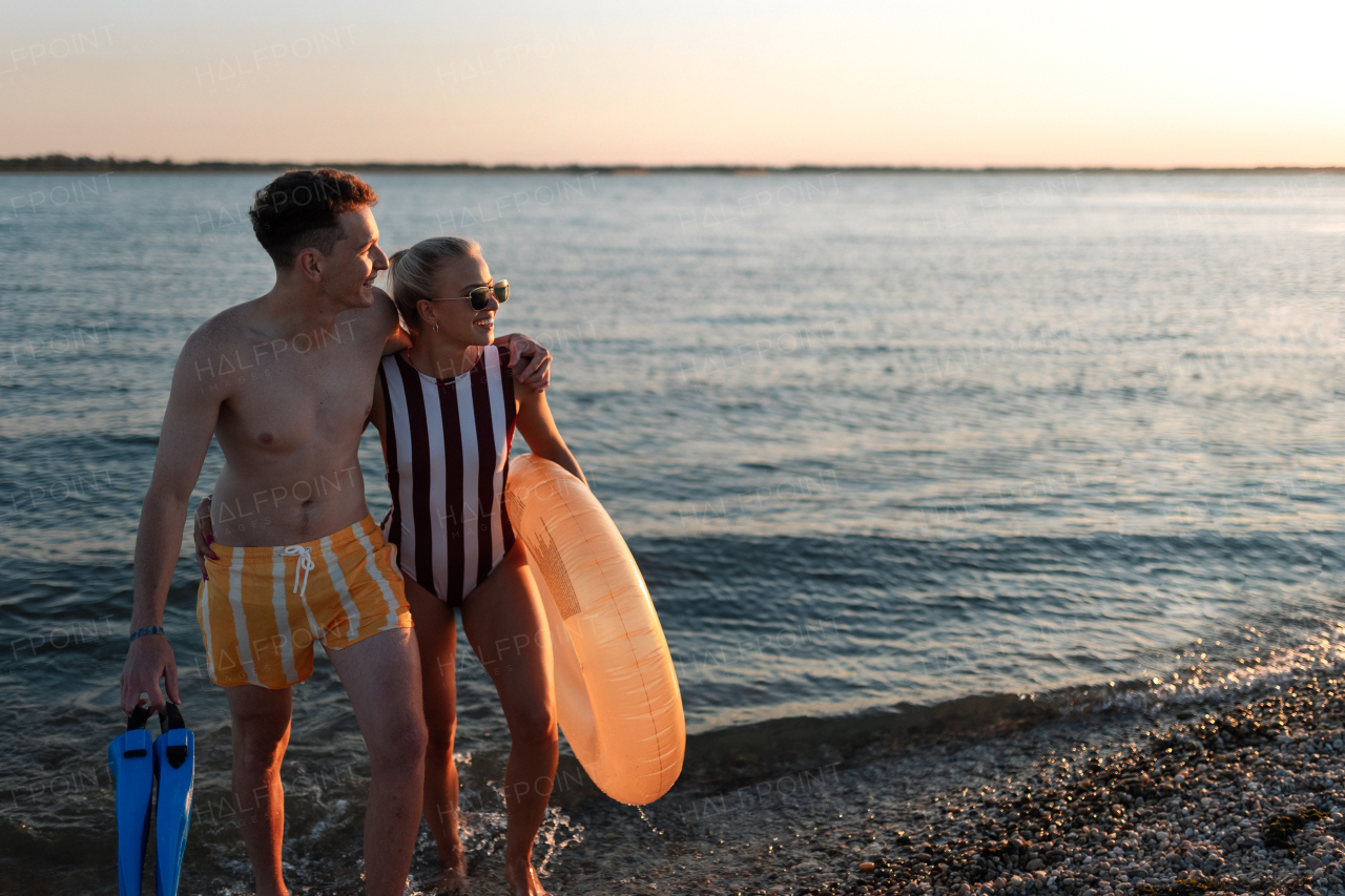 A happy young couple in swimsuits posing with swimming wheel, at beach during sunset.