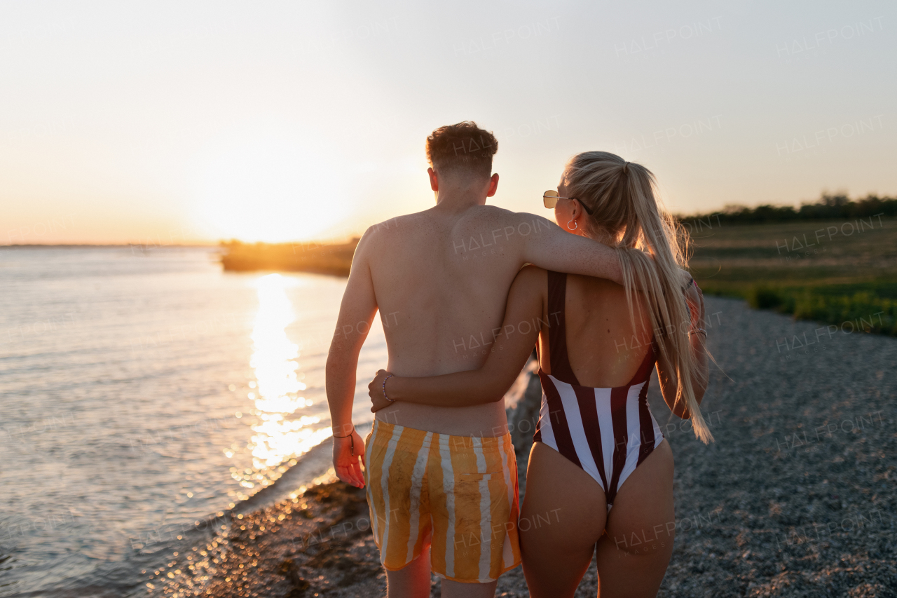 A rear view of young couple in striped swimsuits walking in the beach during sunset.