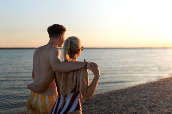 A rear view of young couple in striped swimsuits walking in the beach during sunset.