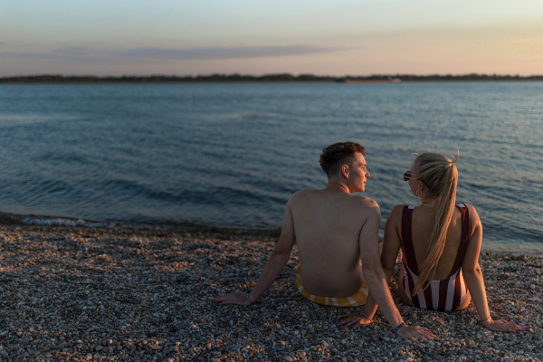 A rear view of young couple in striped swimsuits sitting on the beach during sunset.