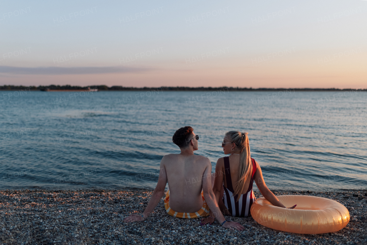 A rear view of young couple in striped swimsuits sitting on the beach during sunset.