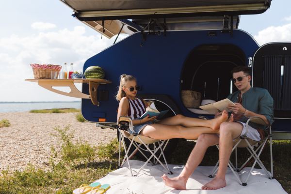 Happy young couple sitting together in front of van, camping and reading a book.