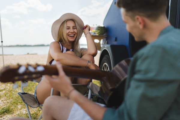 Happy young couple sitting together in front of van, camping and playing at a guitair.