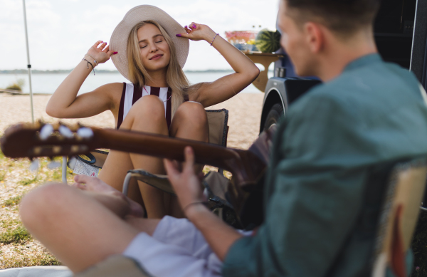 Happy young couple sitting together in front of van, camping and playing at a guitair.