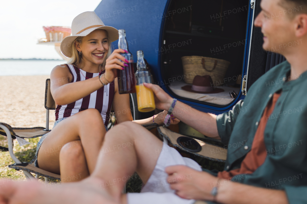 Happy young couple sitting together in front of van, camping and having toast with bottled beer.