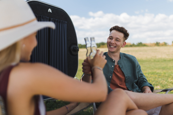 Happy young couple sitting together in front of van, camping and having toast with bottled beer.