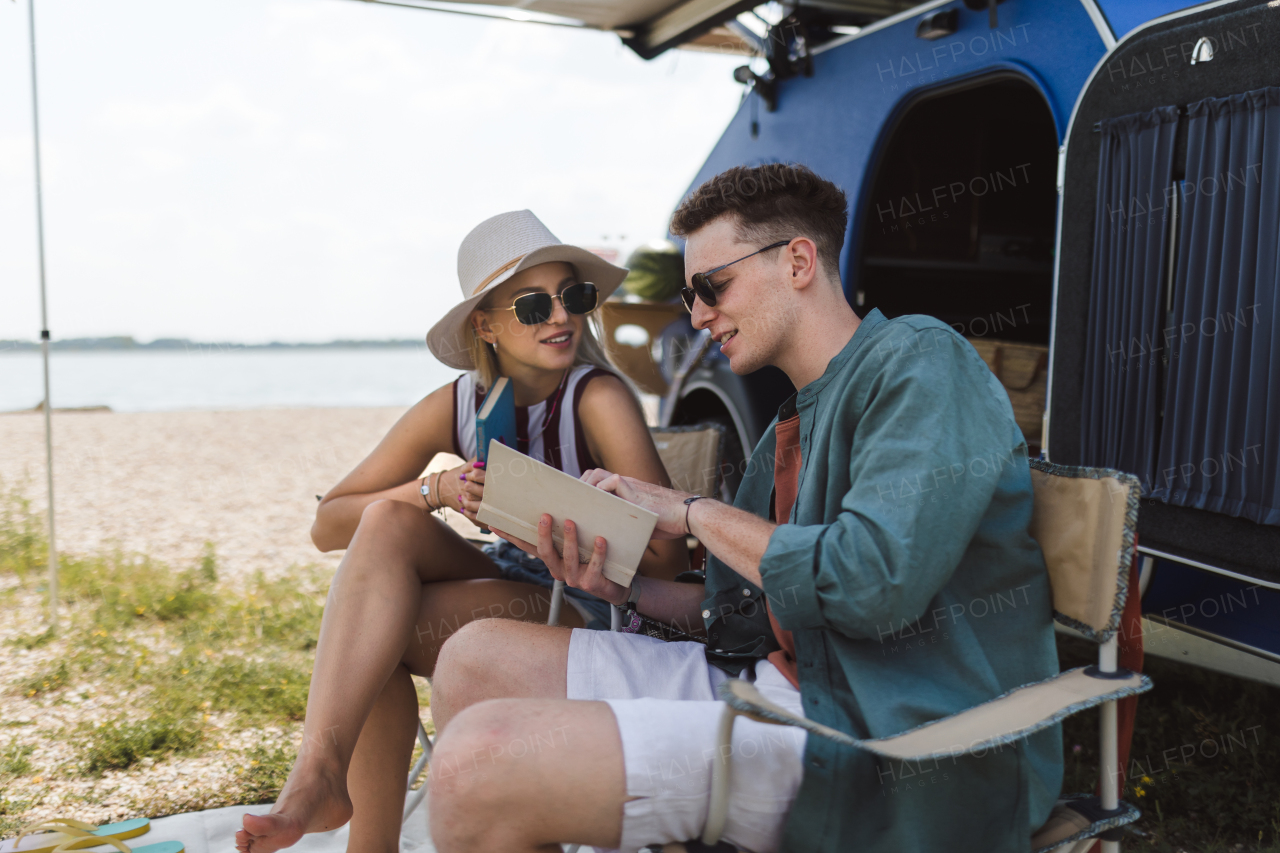 Happy young couple sitting together in front of van, camping and reading a book.