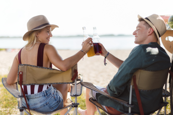 Happy young couple sitting together in front of van, camping and having toast with bottled beer.