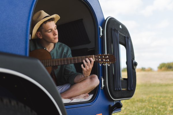 Happy young man sitting in a van, camping ang playing at guitair.