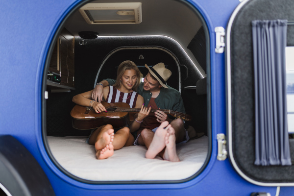 Happy young couple sitting together in a van, camping and playing at a guitair.