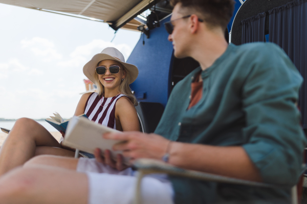 Happy young couple sitting together in front of van, camping and reading a book.