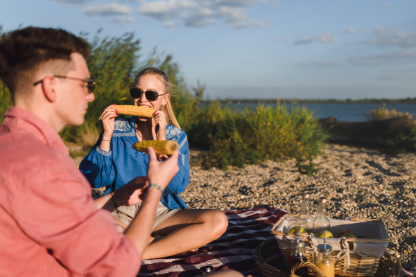 A happy young couple dating together in beach, sitting on a blanket and eating corn. Enjoying holiday time together.