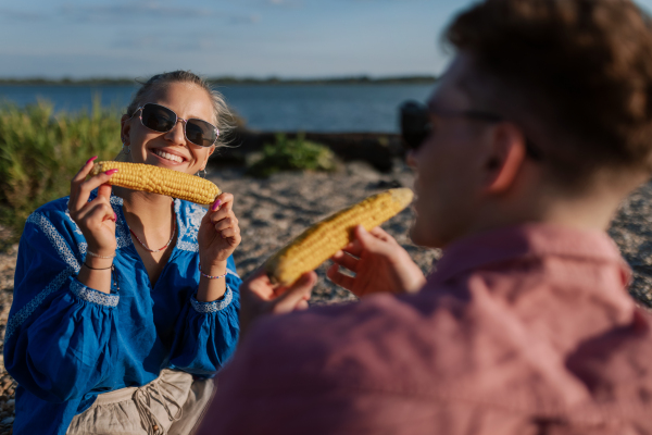 A happy young couple dating together in beach, sitting on a blanket and eating corn. Enjoying holiday time together.