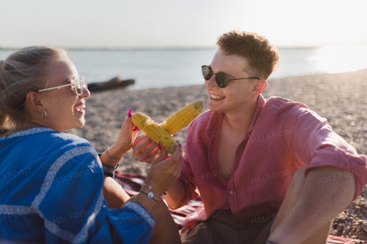 A happy young couple dating together in beach, sitting on a blanket and eating corn. Enjoying holiday time together.