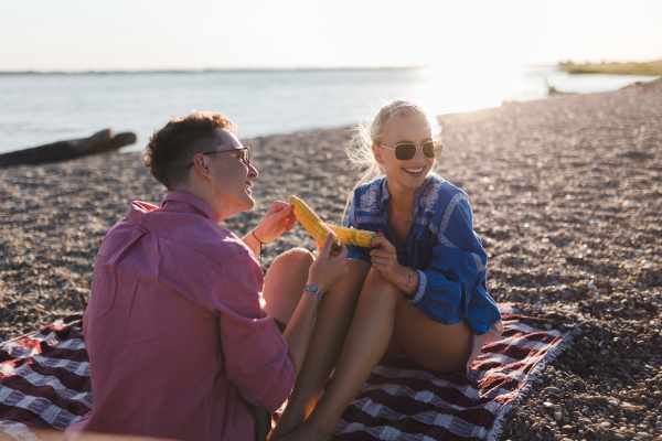 A happy young couple dating together in beach, sitting on a blanket and eating corn. Enjoying holiday time together.