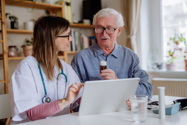 A healthcare worker or caregiver visiting senior man indoors at home, explaining medicine dosage.