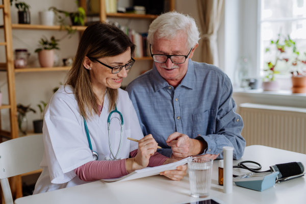 A healthcare worker or caregiver visiting senior man indoors at home, explaining medicine dosage.