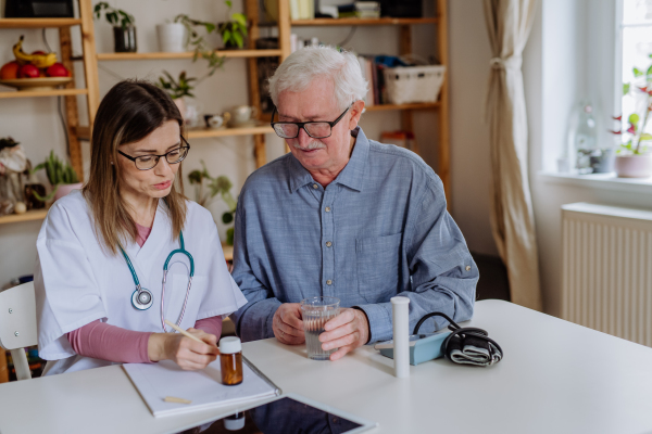 A healthcare worker or caregiver visiting senior man indoors at home, explaining medicine dosage.