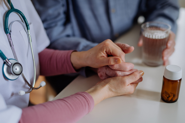 A close-up of doctor holding hand of senior patient and consoling him during medical visit at home.