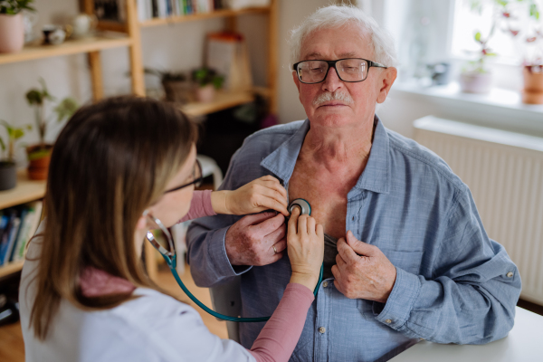 A doctor visiting senior man and examinig him with stethoscope indoors at home, listening to heartbeat.