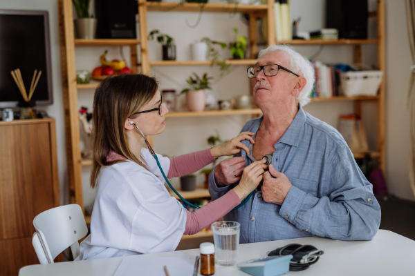 A female doctor visiting senior man and examinig him with stethoscope indoors at home, listening to heartbeat.