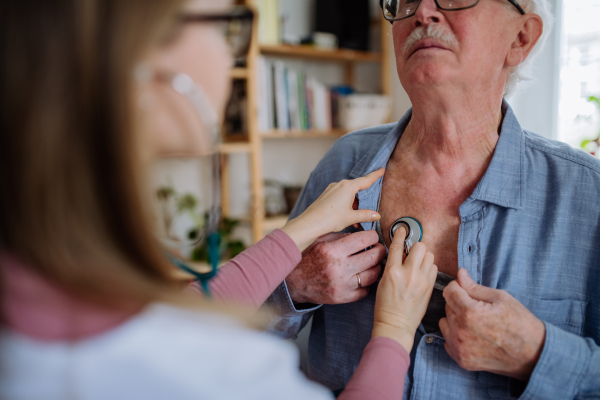 A doctor visiting senior man and examinig him with stethoscope indoors at home, listening to heartbeat.