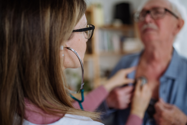 A female doctor visiting senior man and examinig him with stethoscope indoors at home, listening to heartbeat.