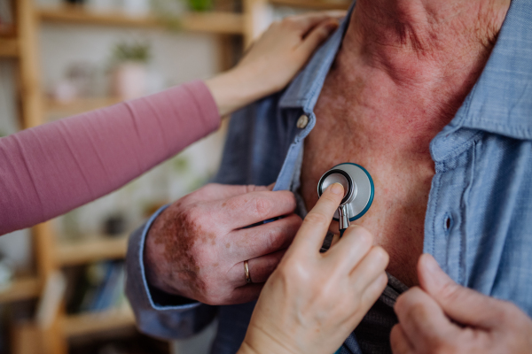 A doctor visiting senior man and examinig him with stethoscope indoors at home, listening to heartbeat.