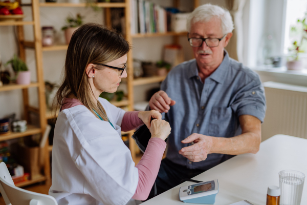 A female doctor visiting senior man and examinig him indoors at home, measuring blood pressure.