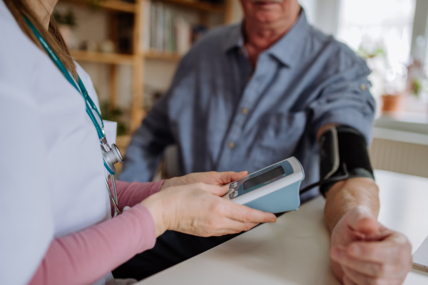 A female doctor visiting senior man and examinig him indoors at home, measuring blood pressure.