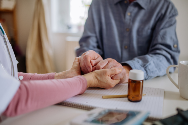 A close-up of doctor holding hand of senior patient and consoling him during medical visit at home.