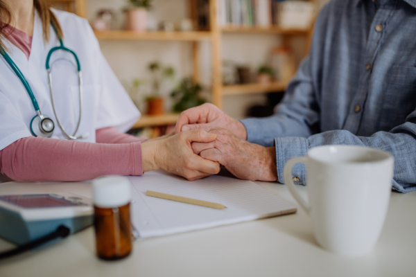 A close-up of doctor holding hand of senior patient and consoling him during medical visit at home.