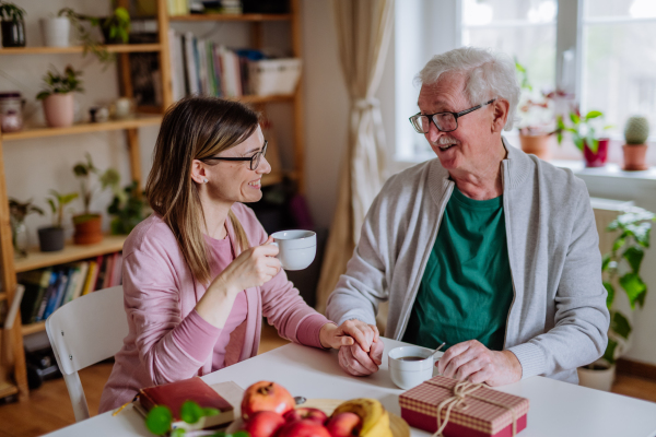 An adult daughter visiting her senior father at home and having coffee together, touching hand.