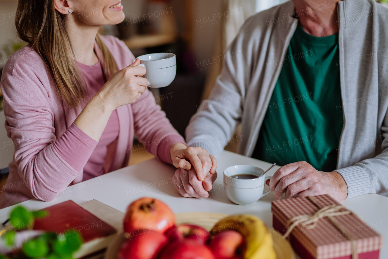 An adult daughter visiting her senior father at home and having coffee together, touching hand.