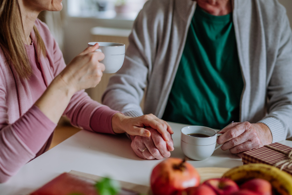 An adult daughter visiting her senior father at home and having coffee together, touching hand.